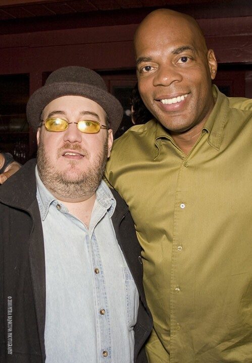 Alonzo Bodden and Brian Fischler standing shoulder to shoulder in a dimly lit club, both smiling toward the camera. Bodden, on the right, is tall and clean-shaven with a broad grin, wearing an olive green button-down shirt. Brian wears a brown felt hat, yellow-tinted glasses, a denim shirt and a dark jacket.