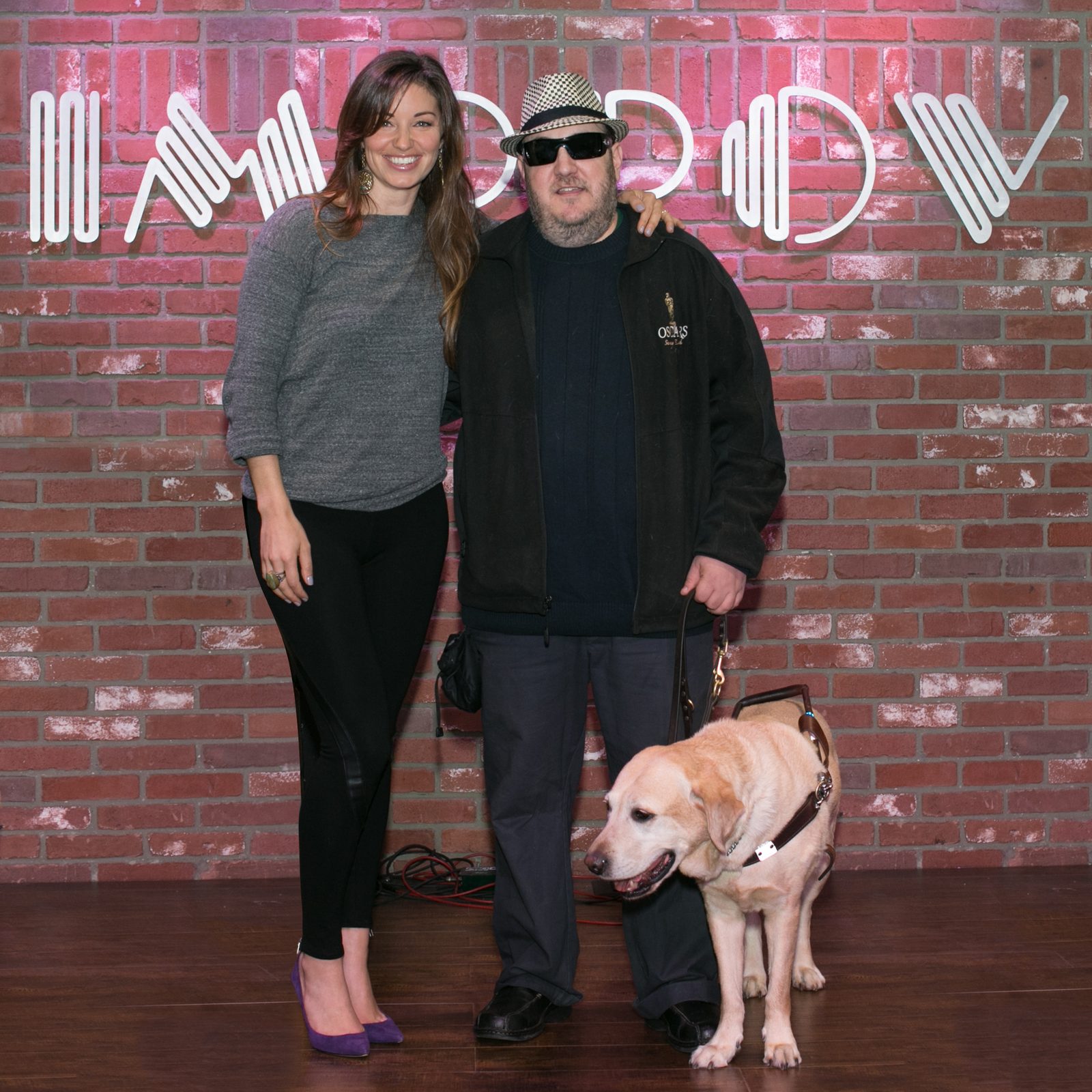 Bianca Kajlich and Brian Fischler standing against an exposed brick wall beneath the Improv's white neon sign. Kajlich has long brown hair and wears a gray sweater, black leggings and purple suede heels, one hand on Brian's shoulder. Brian wears a black and white houndstooth fedora, dark sunglasses and a black jacket embroidered with an Oscars statuette, holding a leather harness handle. Nash, his yellow Labrador guide dog, stands in harness at his feet with his head lowered, the fur around his muzzle gone white.