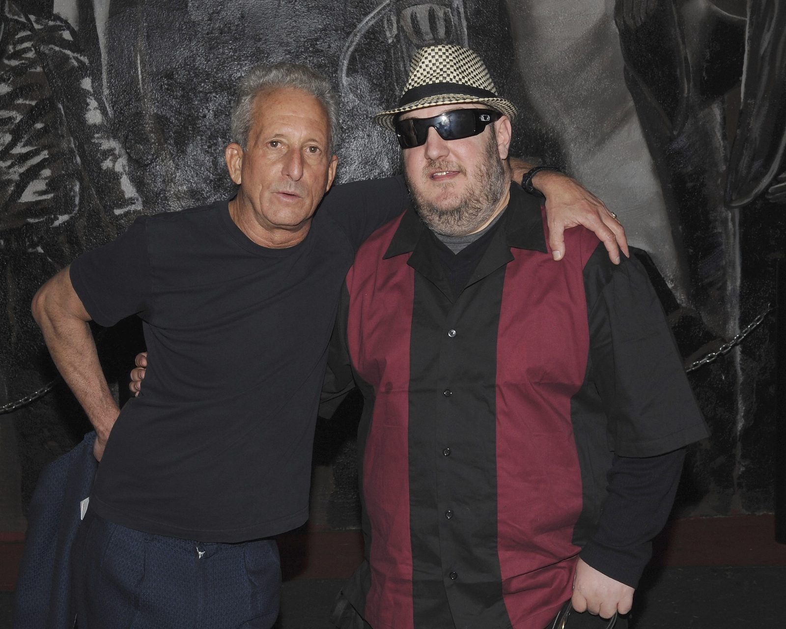 Bobby Slayton and Brian Fischler standing together against a large black and white mural, Brian's arm around Slayton's shoulders. Slayton has short silver-gray hair and wears a plain black T-shirt and navy trousers, one hand on his hip. Brian wears a black and white houndstooth fedora, dark wraparound sunglasses and a maroon and black paneled bowling shirt over a gray tee.