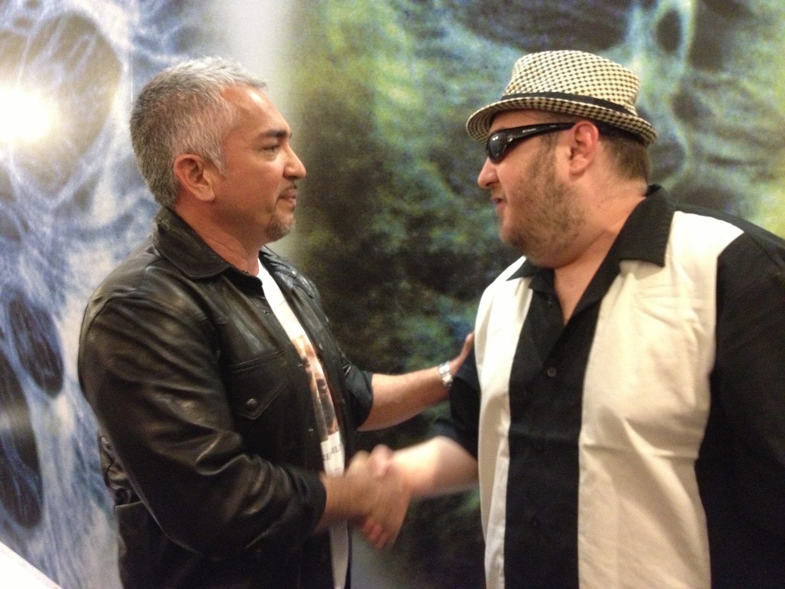 Cesar Millan and Brian Fischler shaking hands and facing each other in profile, Millan's other hand on Brian's shoulder. Millan has short silver hair and a goatee and wears a black leather jacket over a printed T-shirt. Brian wears a black and white houndstooth fedora, dark sunglasses, and a cream and black paneled bowling shirt. A large photographic mural fills the wall behind them.
