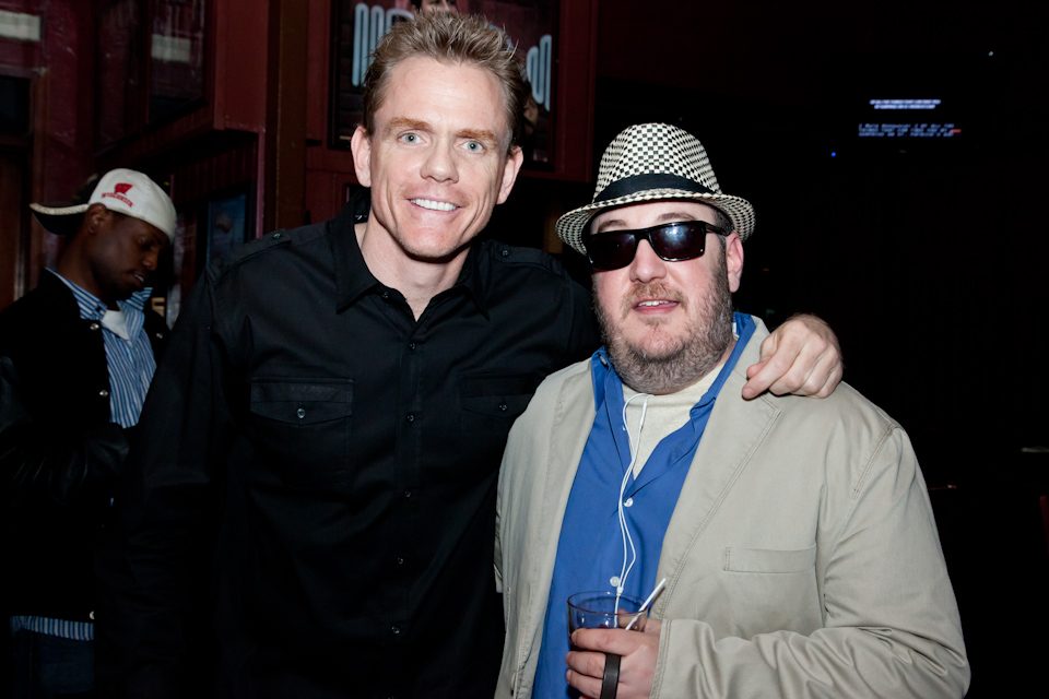 Christopher Titus with his arm around Brian Fischler's shoulders, both smiling at the camera. Titus is tall with short fair hair and wears a black button-down shirt. Brian wears a black and white houndstooth fedora, dark sunglasses, and a tan jacket over a blue shirt, holding a plastic cup with a straw. The darkened club room stretches behind them.