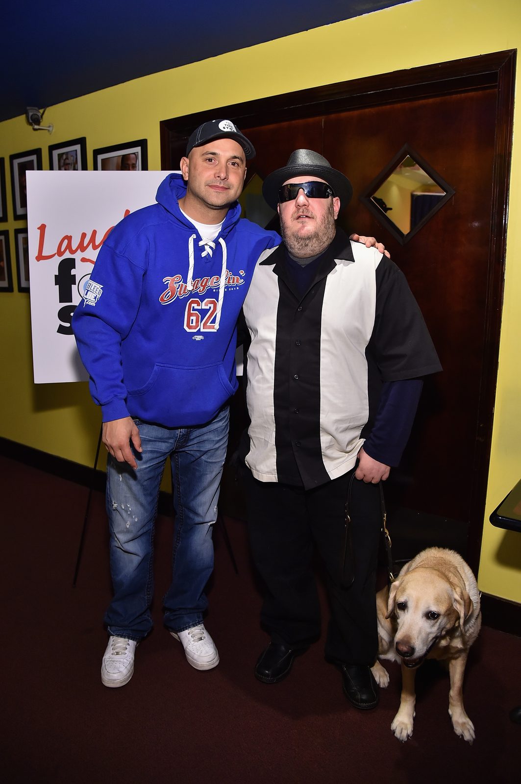 Craig Carton and Brian Fischler standing together in front of the Laugh For Sight sign on its easel, Brian's arm around Carton's shoulders. Carton wears a dark baseball cap and a bright blue lace-neck hooded sweatshirt with the number 62 on the front, over jeans and white sneakers. Brian wears a black straw fedora, dark wraparound sunglasses, and a black and white paneled bowling shirt over a navy long-sleeved top, holding a leather harness handle. Nash, his yellow Labrador guide dog, stands in harness beside him with his head lowered.