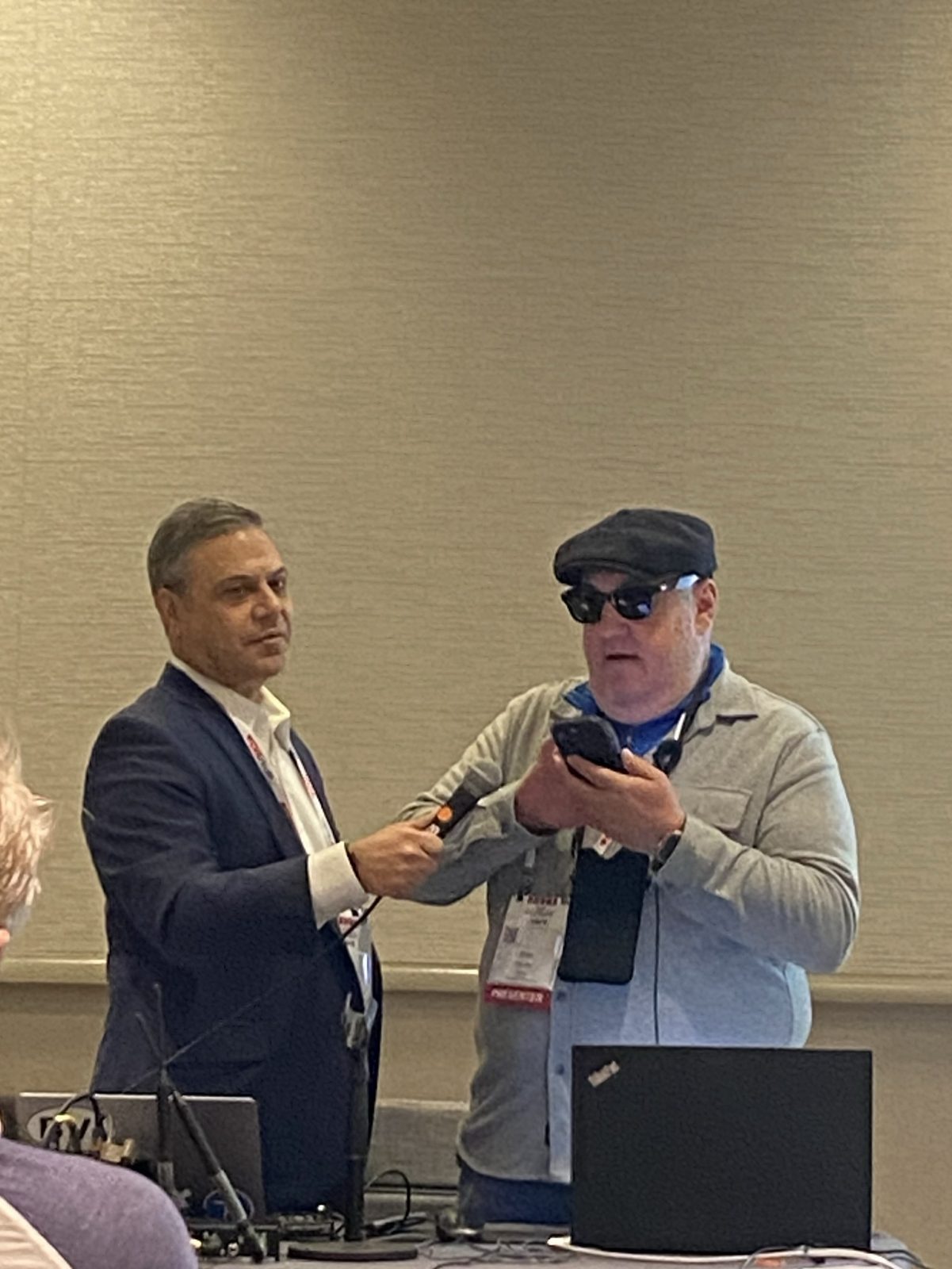 Close view of the same two presenters. Wolynski, in a navy blazer, extends a microphone toward Fischler, who is speaking while holding his phone up in front of him. Fischler wears a black flat cap, dark wraparound sunglasses and a gray zip jacket, with a presenter badge on a lanyard. A black laptop sits on the table in front of them.