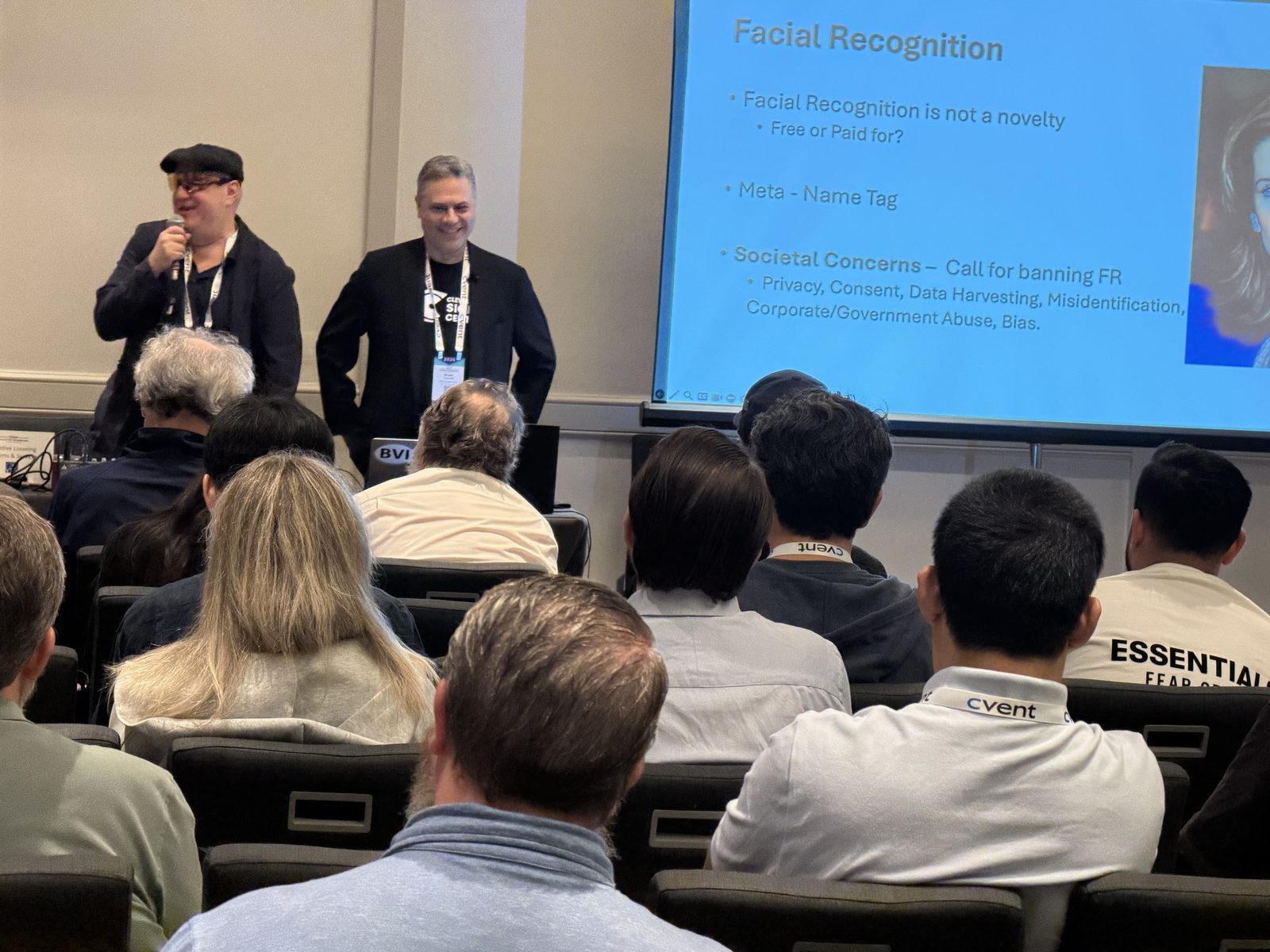 Fischler and Wolynski at the front of a conference session room. Fischler, on the left, wears a dark flat cap, mirrored wraparound sunglasses and a dark jacket, and is speaking into a handheld microphone. Wolynski stands to his right in a black jacket over a dark T-shirt, smiling, a conference lanyard around his neck. A large projection screen fills the right of the frame and the backs of a seated audience fill the foreground.