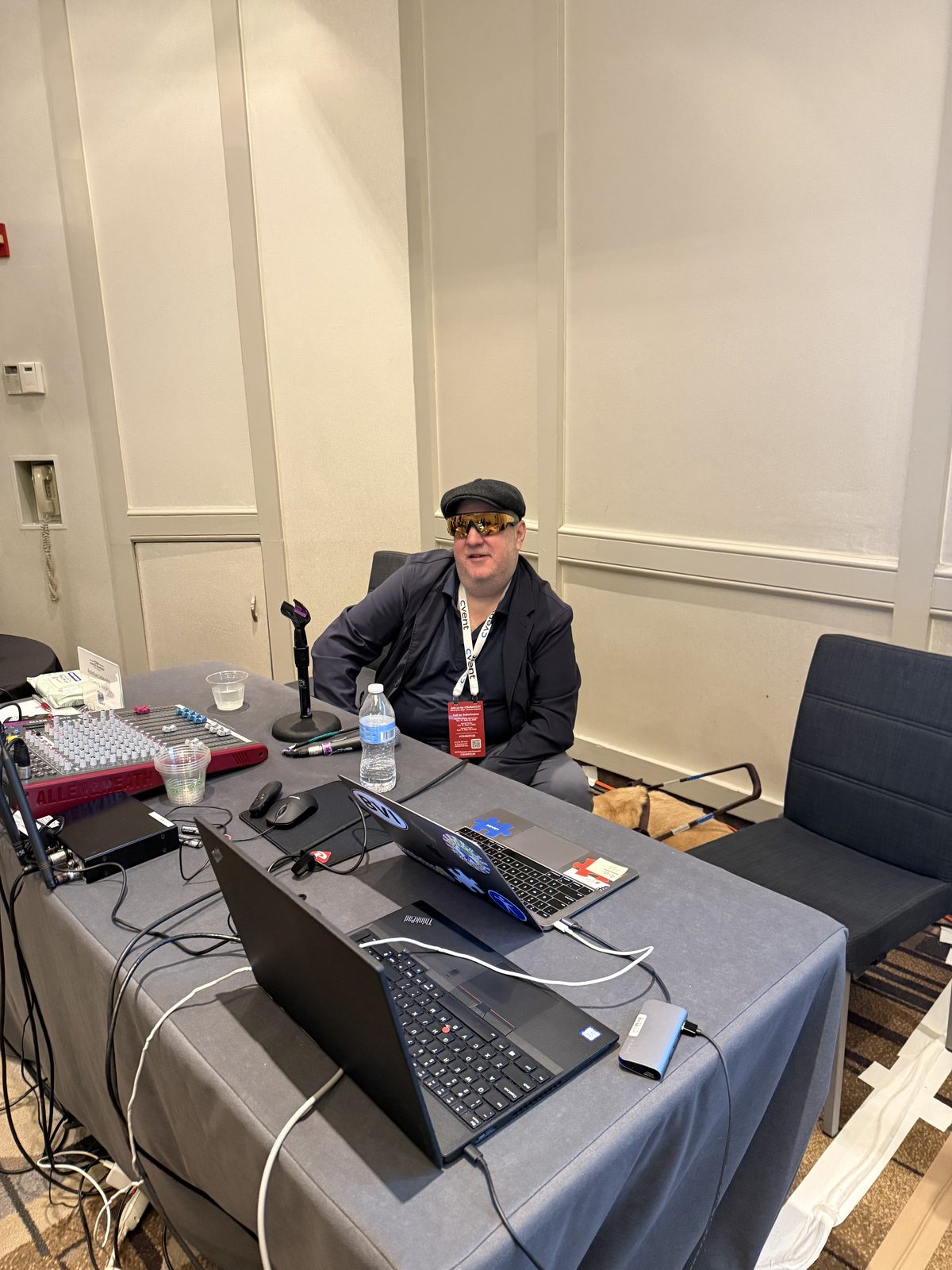 Fischler seated at the presenter table in a conference room, wearing a dark flat cap, mirrored wraparound sunglasses and a dark jacket, with a red conference badge on a lanyard. The table in front of him holds an audio mixer, a microphone on a stand, two open laptops, a bottle of water and a run of cables. His guide dog lies on the floor under the table beside him.