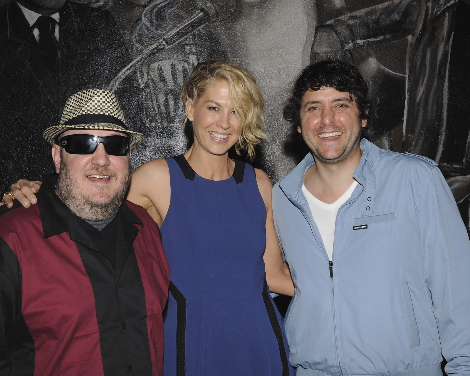 Brian Fischler, Jenna Elfman and Judah Friedlander standing together against a large black and white mural. Brian, on the left, wears a black and white houndstooth fedora, dark wraparound sunglasses and a dark red bowling shirt with a black collar. Elfman, in the center, wears a blue sleeveless dress with black trim, an arm around each of them. Friedlander, on the right, has dark curly hair and wears a pale blue zip-up jacket over a white T-shirt.