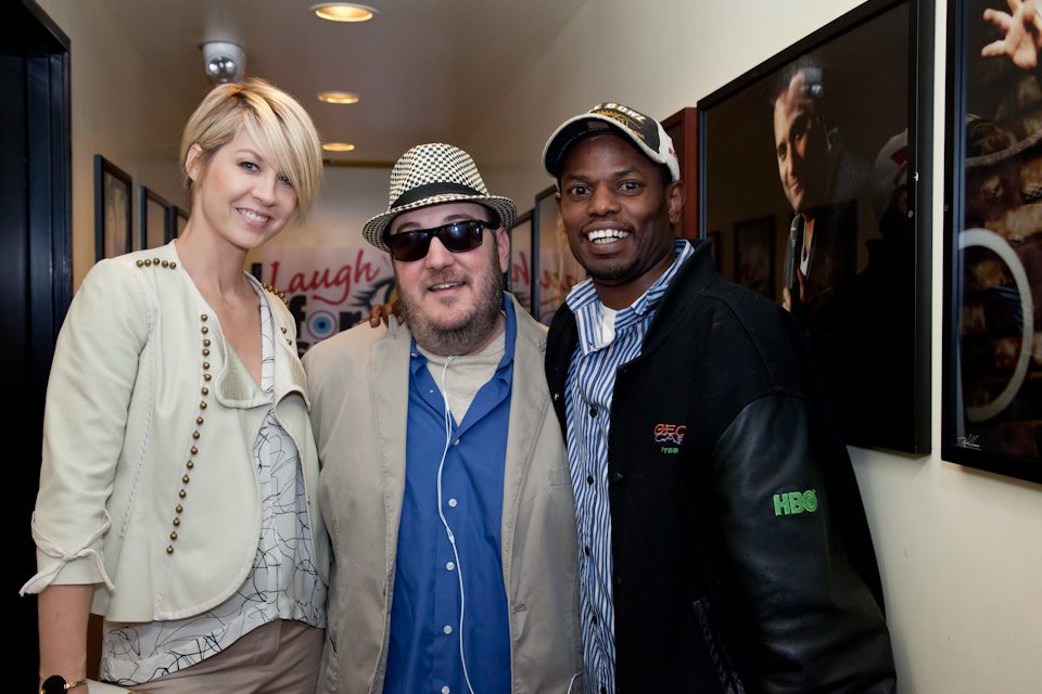 Jenna Elfman, Brian Fischler and Daryl Wright standing together in front of a Laugh For Sight banner. Elfman, on the left, has short blonde hair and wears a cream jacket studded with metal over a patterned blouse. Brian, in the center, wears a black and white houndstooth fedora, dark sunglasses, and a tan jacket over a blue shirt. Wright, on the right, wears a dark cap, a blue and white striped shirt, and a black jacket with leather sleeves carrying an HBO logo. Framed photographs line the wall beside them.