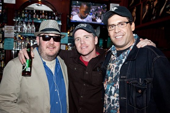 Three men standing together in front of a bar lined with bottles. On the left, Brian Fischler in a black and white houndstooth fedora, dark sunglasses, and a tan jacket over a blue shirt, holding a green beer bottle. In the center, Greg Fitzsimmons in a dark cap and a deep red button-down shirt. On the right, Pablo Francisco in a black cap, wire-framed glasses, and a denim jacket over a shirt printed with photographs. A television screen glows on the wall behind them.
