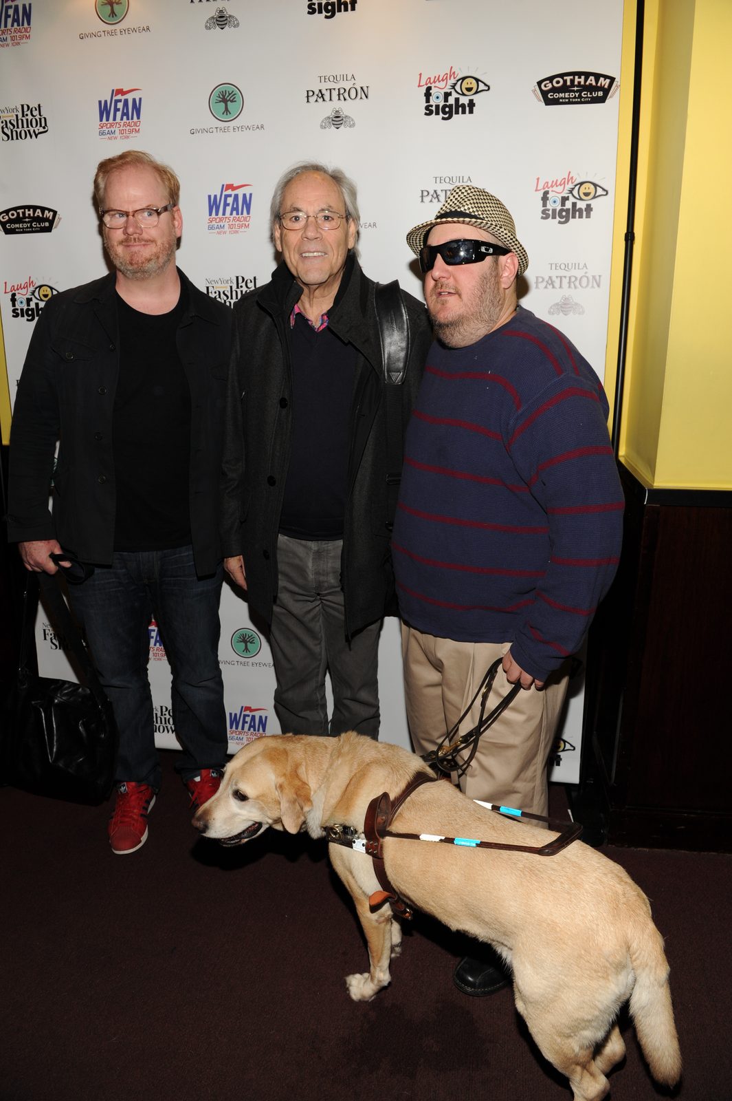 Jim Gaffigan, Robert Klein and Brian Fischler standing together in front of a Laugh For Sight sponsor backdrop, with Brian's yellow Labrador guide dog Nash in harness in front of them.