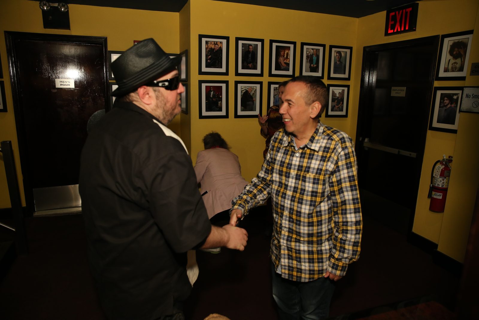 Gilbert Gottfried and Brian Fischler shaking hands in a club corridor with yellow walls hung with framed photographs of comedians. Gottfried, on the right, wears a yellow, blue and white plaid shirt over jeans and is smiling. Brian, on the left, is seen from behind and in profile, in a black straw fedora, dark sunglasses and a black and white paneled bowling shirt. A lit exit sign glows above them.