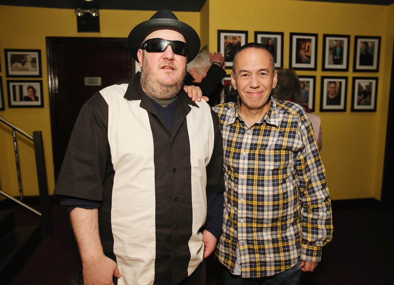 Gilbert Gottfried standing beside Brian Fischler with a hand on his shoulder, both facing the camera. Gottfried wears a yellow, blue and white plaid shirt. Brian wears a black straw fedora, dark wraparound sunglasses, and a black and white paneled bowling shirt over a navy long-sleeved top. Rows of framed photographs of comedians cover the yellow wall behind them.
