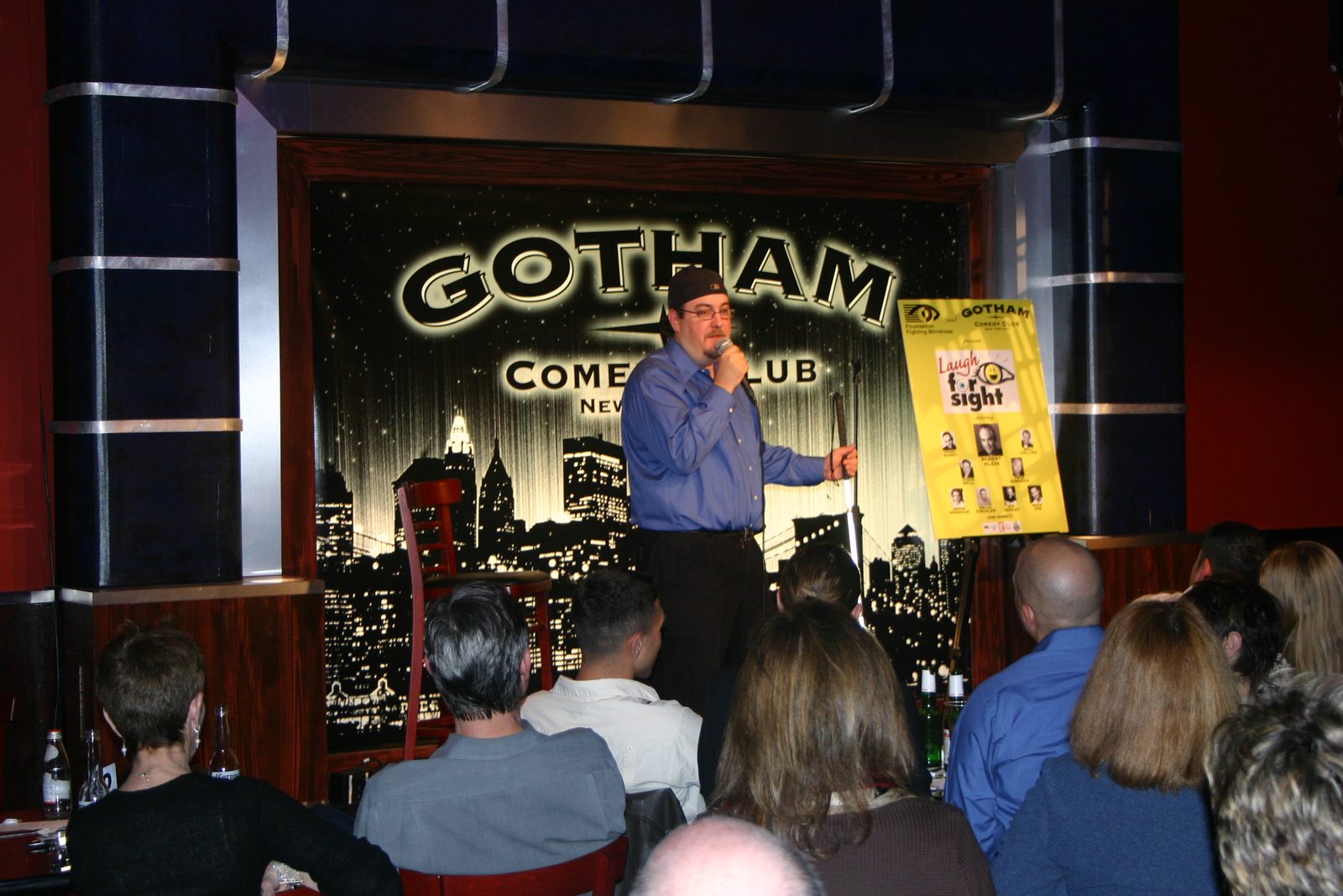 Brian Fischler on stage at Gotham Comedy Club, in a black knit cap, glasses and a blue button-down shirt, holding a microphone to his mouth with one hand and a folded white cane in the other. Behind him the club's backdrop shows the Gotham Comedy Club name in gold above a black and white New York skyline. To his right, an easel holds a yellow poster announcing Laugh For Sight, presented by the Foundation Fighting Blindness and Gotham Comedy Club, with Robert Klein billed at the center above headshots of the rest of the lineup. The backs of the seated audience fill the foreground.