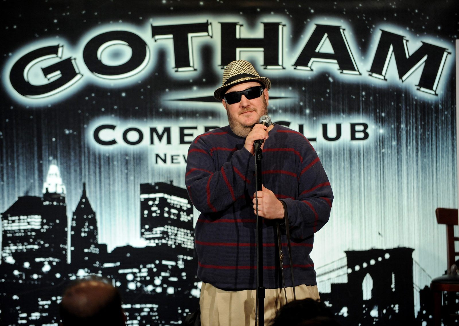 Brian Fischler on stage at Gotham Comedy Club, both hands on the microphone and its stand, head tilted slightly and mid-line. He wears a black and white houndstooth fedora, dark sunglasses, a navy waffle-knit sweater with red horizontal stripes, and khaki trousers. A leather guide dog harness handle hangs from his wrist. The club's backdrop fills the frame behind him, the Gotham Comedy Club name glowing above an illuminated New York skyline.