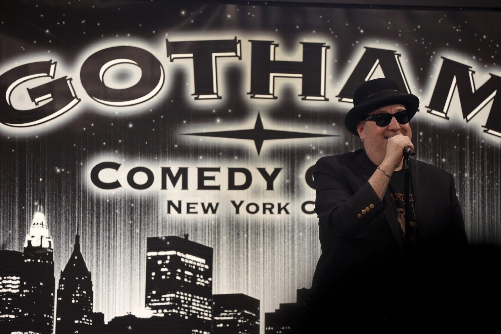 Brian Fischler performing stand-up at Gotham Comedy Club in a black bowler hat and dark sunglasses, one hand on the microphone, the club's lit backdrop behind him.
