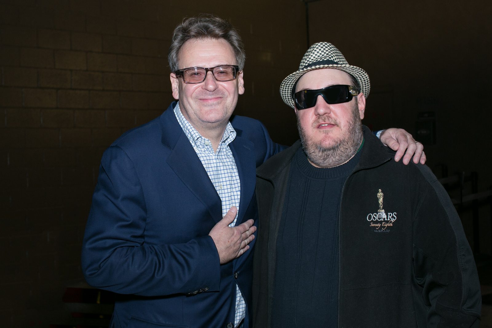 Greg Proops and Brian Fischler standing together against a dark brick wall backstage. Proops wears heavy tortoiseshell glasses, a navy blazer and a blue checked shirt, one hand resting on his chest. Brian wears a black and white houndstooth fedora, dark sunglasses, and a black jacket embroidered with an Oscars statuette over a black sweater, with an arm around Proops.