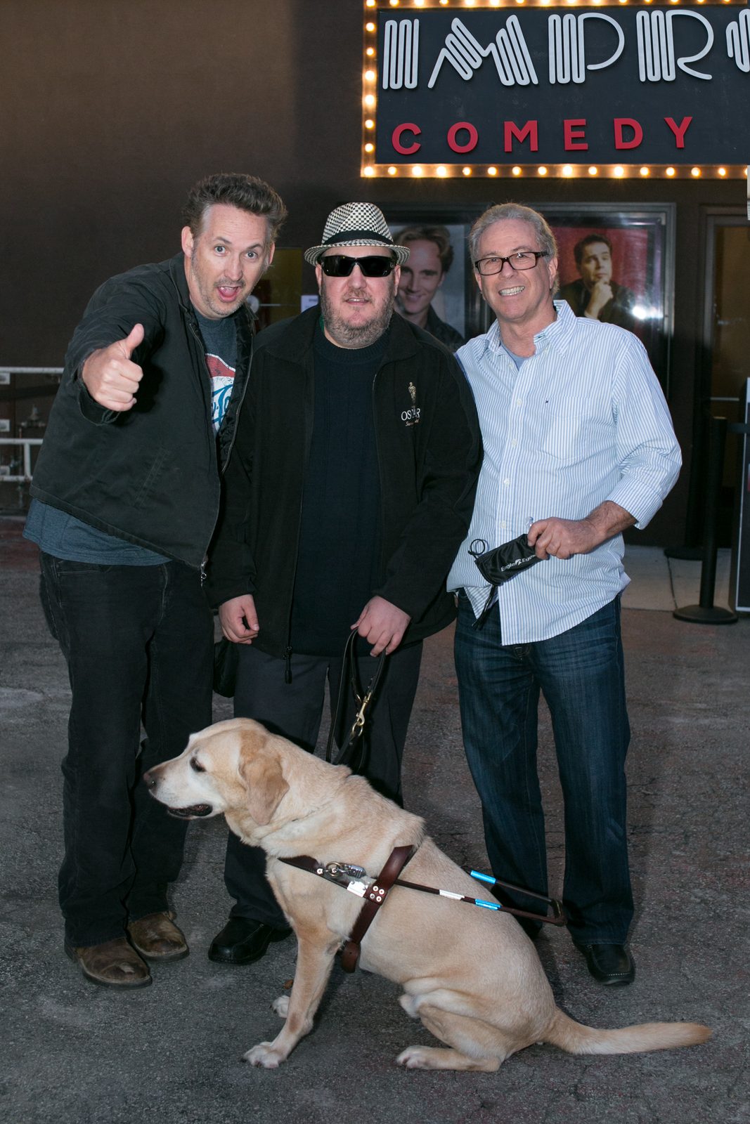 Three men standing outside a comedy club at dusk, beneath an illuminated marquee spelling Improv Comedy in bulbs. On the left, Harland Williams giving a thumbs up with a wide-eyed grin, in a dark jacket over a graphic T-shirt. In the center, Brian Fischler in a black and white houndstooth fedora, dark sunglasses and a black jacket embroidered with an Oscars statuette, holding a guide dog harness. On the right, Bobby Slayton, silver-haired and in dark rectangular glasses, wearing a blue and white striped shirt open over a light tee, holding a small black sunglasses pouch. Brian's yellow Labrador guide dog Nash sits in harness in front of them.