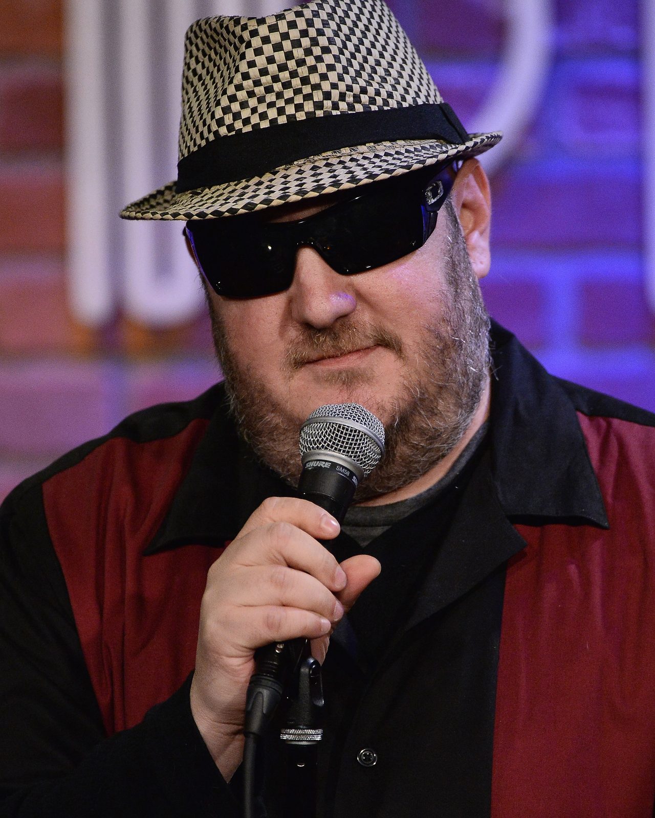 Brian Fischler performing at the Hollywood Improv in a checked trilby and dark wraparound sunglasses, close up on the microphone, mid-line and half-smiling under purple stage light.