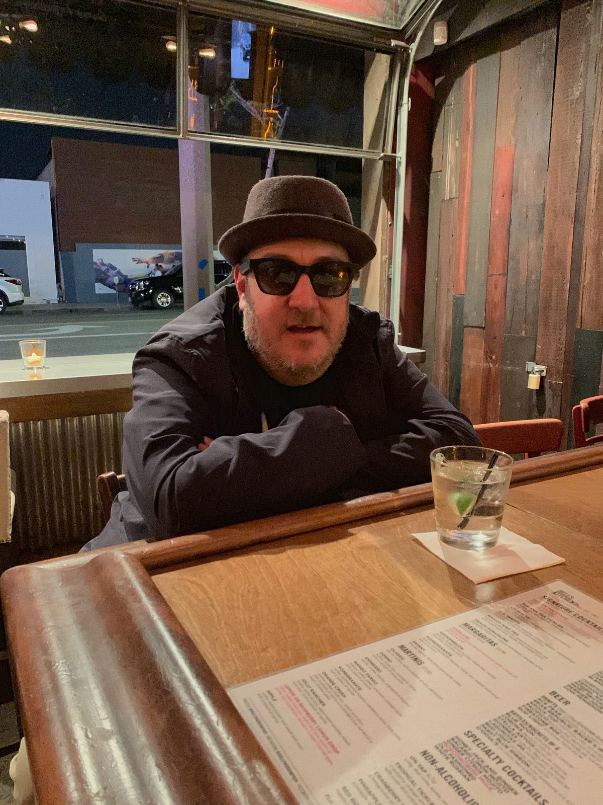 Brian Fischler at the Hollywood Improv at night, in dark sunglasses and a brown felt hat, leaning forward with his arms folded on a wooden counter beside a drink and a cocktail menu.