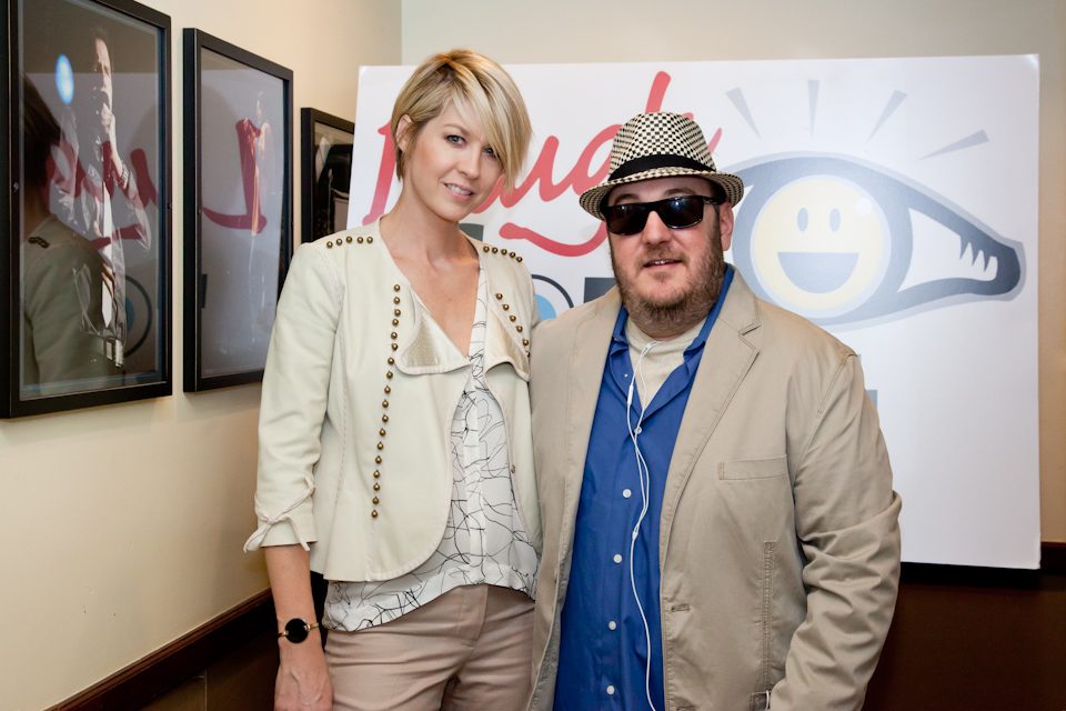 Jenna Elfman and Brian Fischler standing in front of a large Laugh For Sight banner showing the smiling eye logo. Elfman has short blonde hair and wears a cream jacket studded with metal along the lapels over a patterned blouse, with light trousers. Brian wears a black and white houndstooth fedora, dark sunglasses, and a tan jacket over a blue shirt, with white earbuds hanging around his neck.