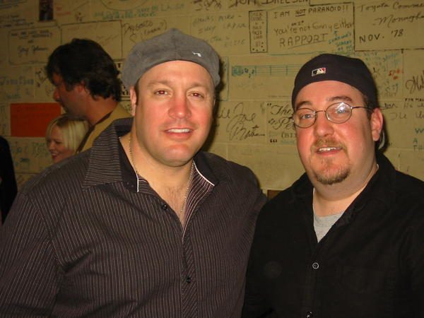 Brian Fischler and Kevin James standing together in front of a wall covered in comedians' signatures and scrawled messages.
