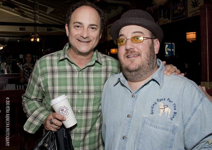 Kevin Pollak with his arm around Brian Fischler's shoulders, both smiling at the camera. Pollak wears a green and white plaid shirt and holds a white paper coffee cup. Brian wears a brown felt hat, yellow-tinted glasses, and a denim shirt embroidered with the Guiding Eyes for the Blind logo. A bar and warm club lighting fill the background.