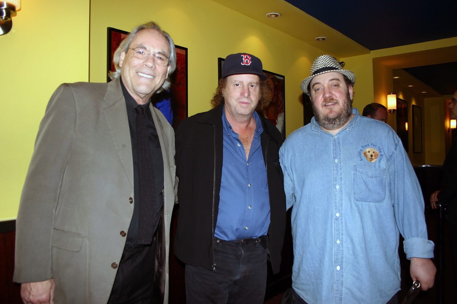 Three men standing together in a club lobby with yellow walls and framed photographs. On the left, Robert Klein, gray-haired and in wire-framed glasses, wearing a tan overcoat over a black shirt and dark tie. In the center, Steven Wright, curly-haired under a navy Boston Red Sox cap, in a blue shirt open at the collar under a black jacket. On the right, Brian Fischler in a black and white houndstooth fedora and a denim shirt embroidered with the Guiding Eyes for the Blind logo.