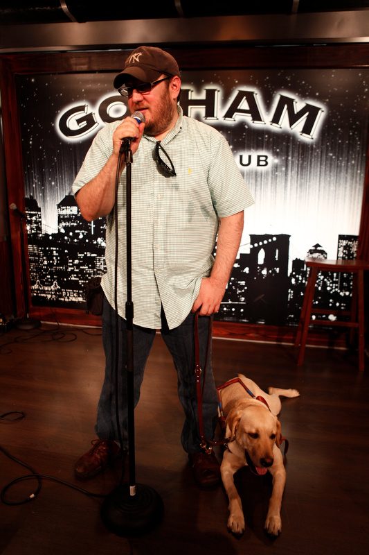 Brian Fischler on stage at Gotham Comedy Club, singing or speaking into a microphone on a stand, one hand on the mic. He wears a brown New York Yankees cap, dark glasses, a mint green checked short-sleeved shirt with sunglasses hooked in the collar, and jeans. In his other hand he holds the leash of his yellow Labrador guide dog Nash, who lies on the stage beside him in harness, mouth open. The club's lit skyline backdrop is behind them.