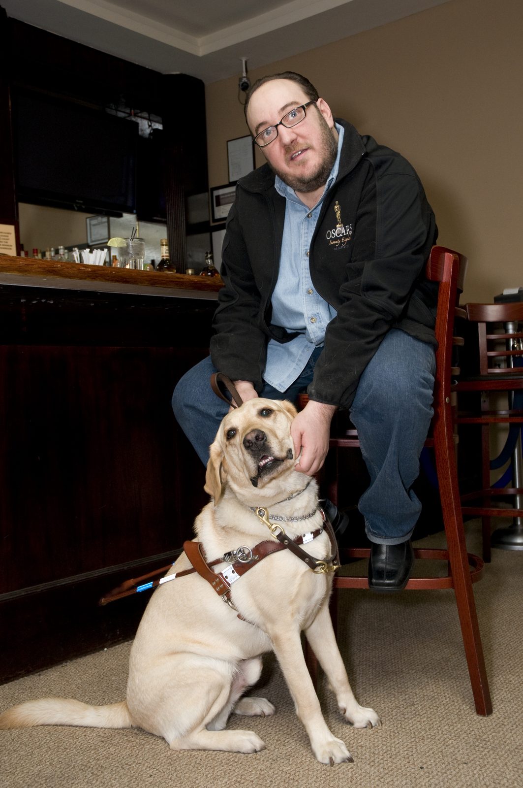 Brian Fischler seated on a tall chair at a bar, in glasses and a beard, wearing a black jacket embroidered with an Oscars statuette over a light blue button-down shirt and jeans. He rests one hand on the head of a yellow Labrador sitting in front of him in a leather guide dog harness, mouth open, looking up and away. A folded white cane rests against the dog's side. Behind them the bar top holds bottles and glasses.