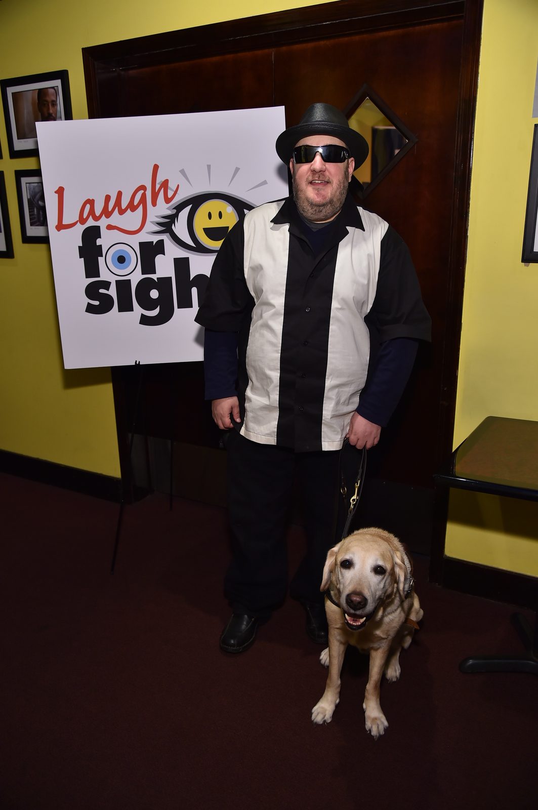 Brian Fischler standing beside a large Laugh For Sight sign propped on an easel, its logo a smiling eye. He wears a black straw fedora, dark wraparound sunglasses, and a black and white paneled bowling shirt over a navy long-sleeved top, holding a leather harness handle. Nash, his yellow Labrador guide dog, sits at his feet looking up at the camera, mouth open. Framed photographs hang on the yellow wall behind them.