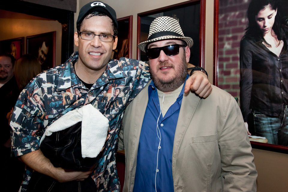 Pablo Francisco with his arm around Brian Fischler. Francisco wears a black cap, wire-framed glasses, and a shirt printed all over with photographs, with a folded coat and a white towel over one arm. Brian wears a black and white houndstooth fedora, dark sunglasses, and a tan jacket over a blue shirt. A large photograph of Sarah Silverman hangs on the brick wall behind them.