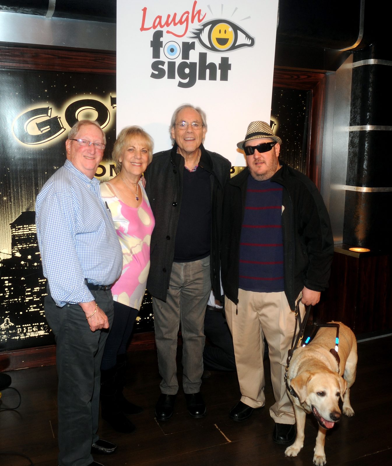 Four people standing in front of a Laugh For Sight banner, the letter O in the word for drawn as a smiling eye, with the lit Gotham Comedy Club skyline backdrop behind. From the left: an older man in a blue checked shirt and glasses, an older woman in a pink and cream patterned top, Robert Klein in a dark overcoat and glasses, and Brian Fischler in a houndstooth fedora and dark sunglasses, wearing a navy sweater with red stripes. Brian's yellow Labrador guide dog Nash stands in harness at his side, mouth open.