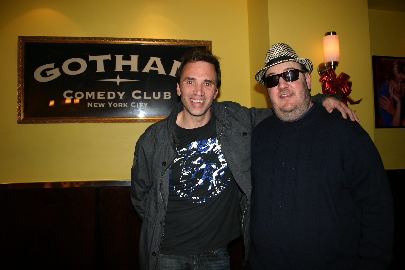 Paul Mercurio and Brian Fischler standing side by side beneath a framed Gotham Comedy Club New York City sign on a yellow wall. Mercurio wears a gray zip-up jacket over a black graphic T-shirt and has his arm around Brian's shoulders. Brian wears a black and white houndstooth fedora, dark sunglasses and a navy sweater. A red holiday bow is pinned to the wall behind them.