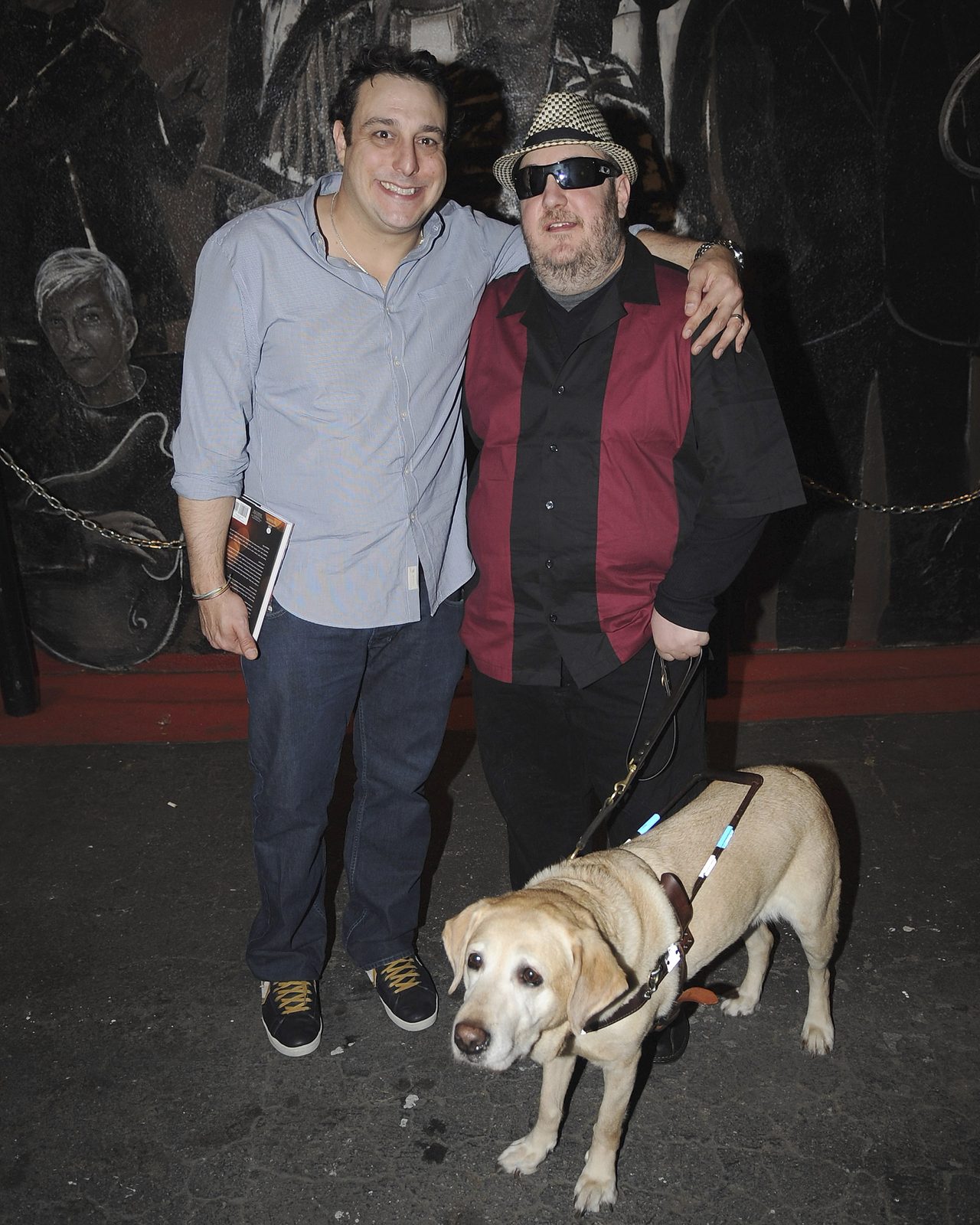 Petros Papadakis and Brian Fischler standing side by side outside the club at night, Brian's arm around his shoulders. Papadakis wears a pale blue button-down shirt with the sleeves rolled and holds a program. Brian wears a black and white houndstooth fedora, dark wraparound sunglasses and a maroon and black paneled bowling shirt, holding a leather harness handle. Nash, his yellow Labrador guide dog, stands in harness at their feet. A large black and white mural covers the wall behind them.