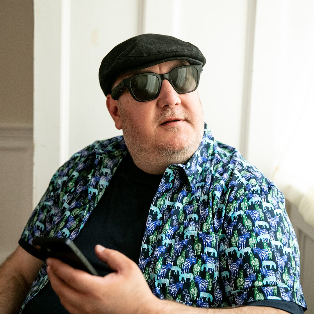 Brian Fischler in dark sunglasses and a black flat cap, wearing a zebra-print shirt over a navy tee, holding his phone by a sunlit window.