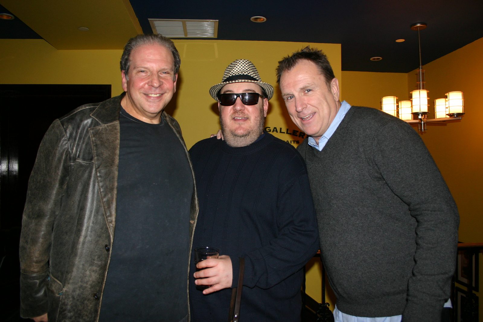 Three men standing together against a yellow wall in a club lobby. On the left, Eddie Brill, heavier-set with graying hair, in a worn brown leather jacket over a dark T-shirt. In the center, Brian Fischler in a black and white houndstooth fedora, dark sunglasses and a navy cable-knit sweater, holding a glass of beer. On the right, Colin Quinn, with short spiked hair, in a charcoal sweater over a pale blue collared shirt, one arm around Brian's shoulders.
