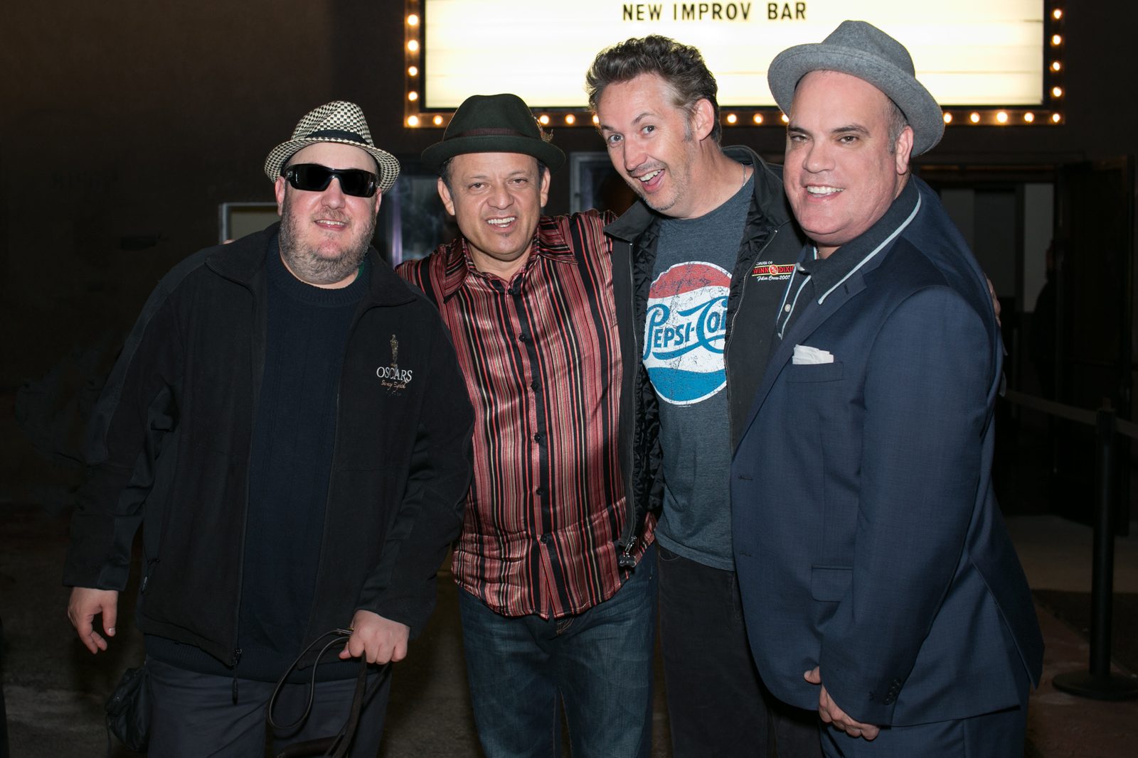 Four men standing together outside the Hollywood Improv, beneath a lit marquee reading New Improv Bar. From the left: Brian Fischler in a black and white houndstooth fedora, dark sunglasses and a black jacket embroidered with an Oscars statuette, holding a guide dog harness; Paul Rodriguez in a black fedora and a red and black striped shirt; Harland Williams, wide-eyed and grinning, in a faded Pepsi-Cola T-shirt under a black jacket; and a fourth man in a gray fedora and a navy suit with a white pocket square.
