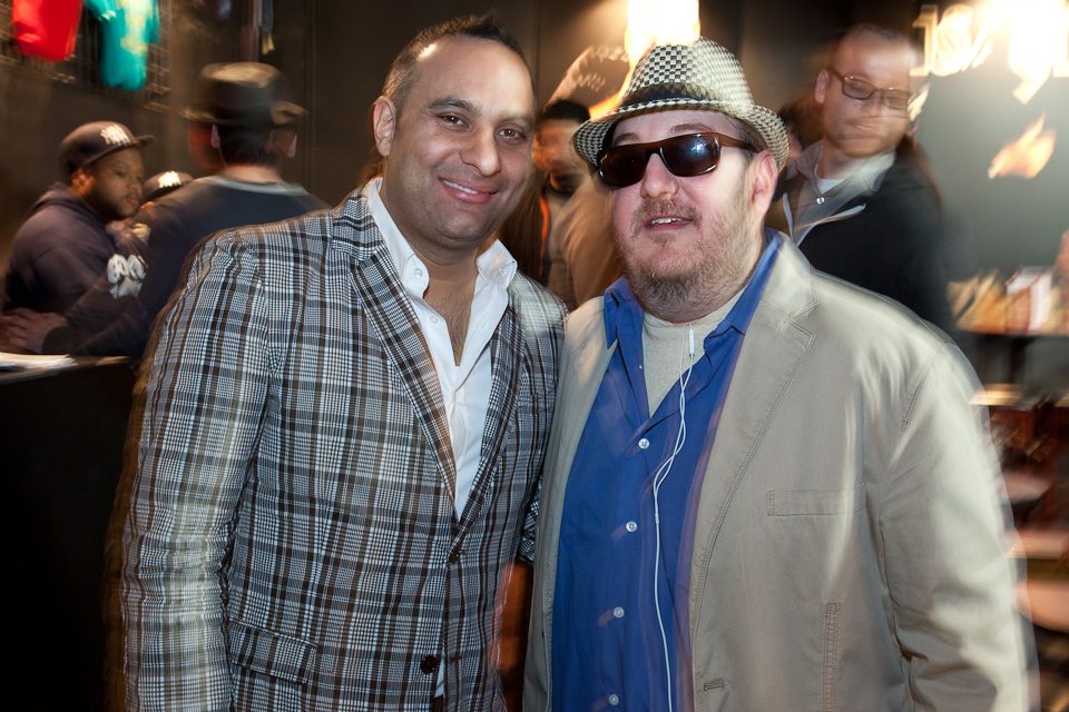 Russell Peters and Brian Fischler standing shoulder to shoulder in a crowded club, both looking at the camera. Peters wears a gray and brown checked blazer over an open-collared white shirt. Brian wears a black and white houndstooth fedora, dark sunglasses, and a tan jacket over a blue shirt with white earbuds around his neck. Other guests fill the room behind them.