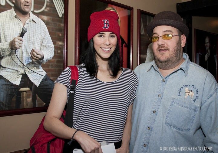 Sarah Silverman and Brian Fischler standing together in a club lobby. Silverman wears a red Boston Red Sox knit cap with the letter B, a navy and white striped top, and a red bag over one shoulder, holding a few sheets of paper. Brian wears a brown felt hat, yellow-tinted glasses, and a denim shirt embroidered with the Guiding Eyes for the Blind logo. Framed photographs of comedians hang on the brick wall behind them.
