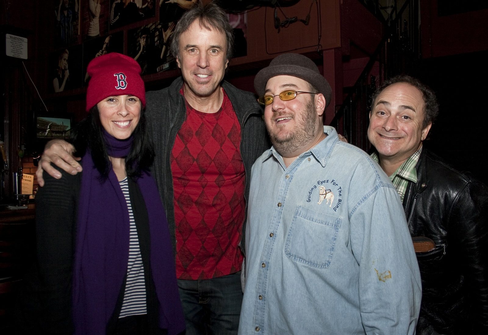 Four people posed together at a bar: Sarah Silverman in a red Boston Red Sox beanie and purple scarf, Kevin Nealon in a red diamond-patterned shirt, Brian Fischler in a brown hat and tinted glasses wearing a denim shirt embroidered with the Guiding Eyes for the Blind logo, and Kevin Pollak in a black leather jacket.