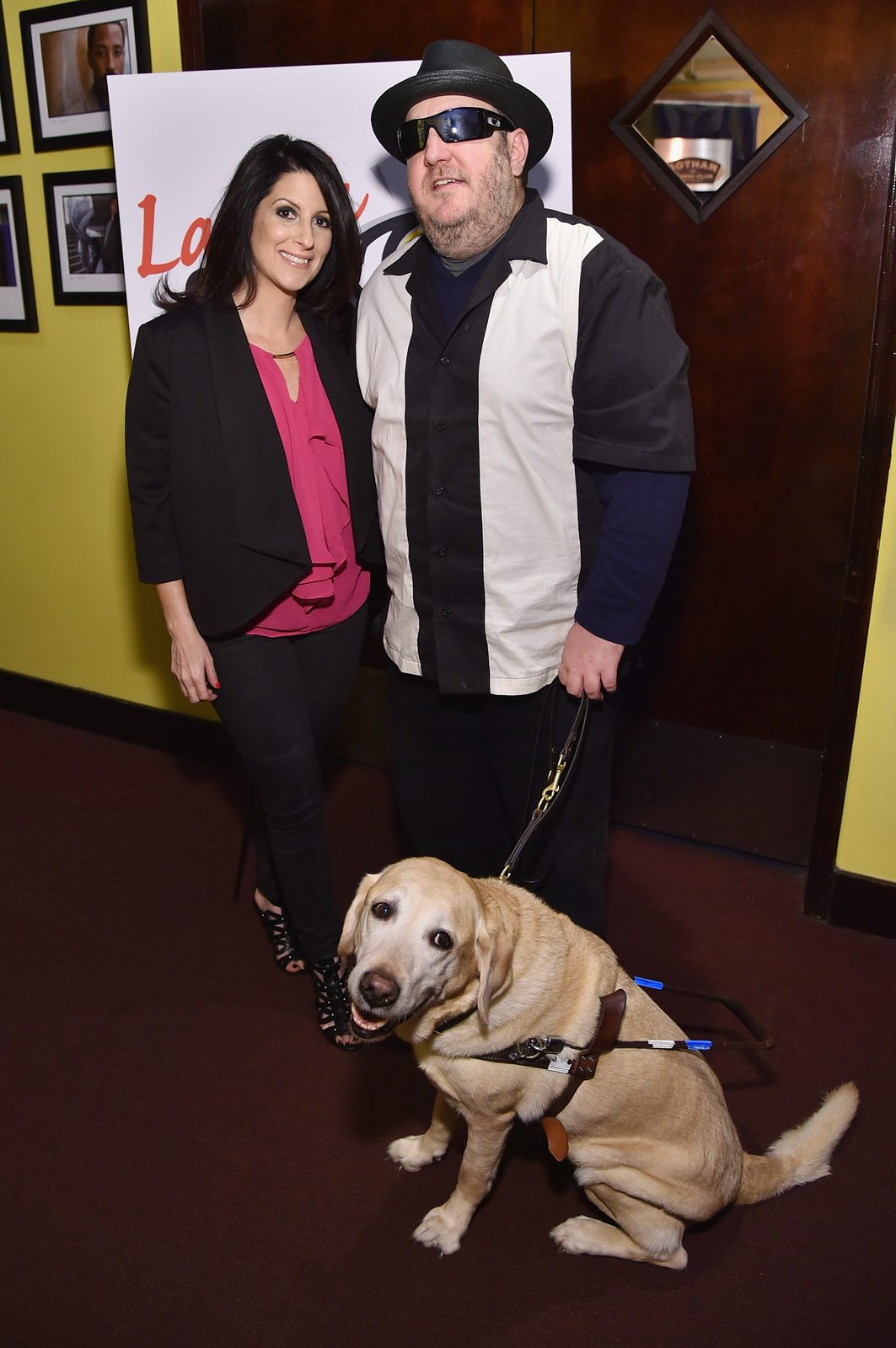 Stacey Prussman and Brian Fischler standing together in front of the Laugh For Sight sign on its easel. Prussman has long dark hair and wears a black blazer over a bright pink blouse with black trousers and strappy heels. Brian wears a black straw fedora, dark wraparound sunglasses, and a black and white paneled bowling shirt over a navy long-sleeved top, holding a leather harness handle. Nash, his yellow Labrador guide dog, sits in harness at their feet, mouth open.