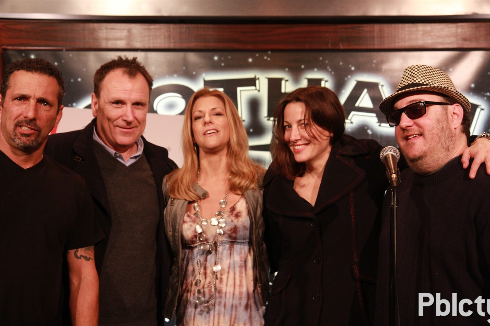 Five people standing shoulder to shoulder on stage in front of the Gotham Comedy Club backdrop, a microphone on a stand at the right of the frame. From the left: Rich Vos in a black T-shirt with a tattoo on his forearm; Colin Quinn in a charcoal sweater over a pale blue collared shirt; a blonde woman in a patterned dress with long layered necklaces and a gray jacket; a dark-haired woman in a black winter coat; and Brian Fischler in a black and white houndstooth fedora, dark sunglasses and a navy sweater, with an arm around her shoulders.