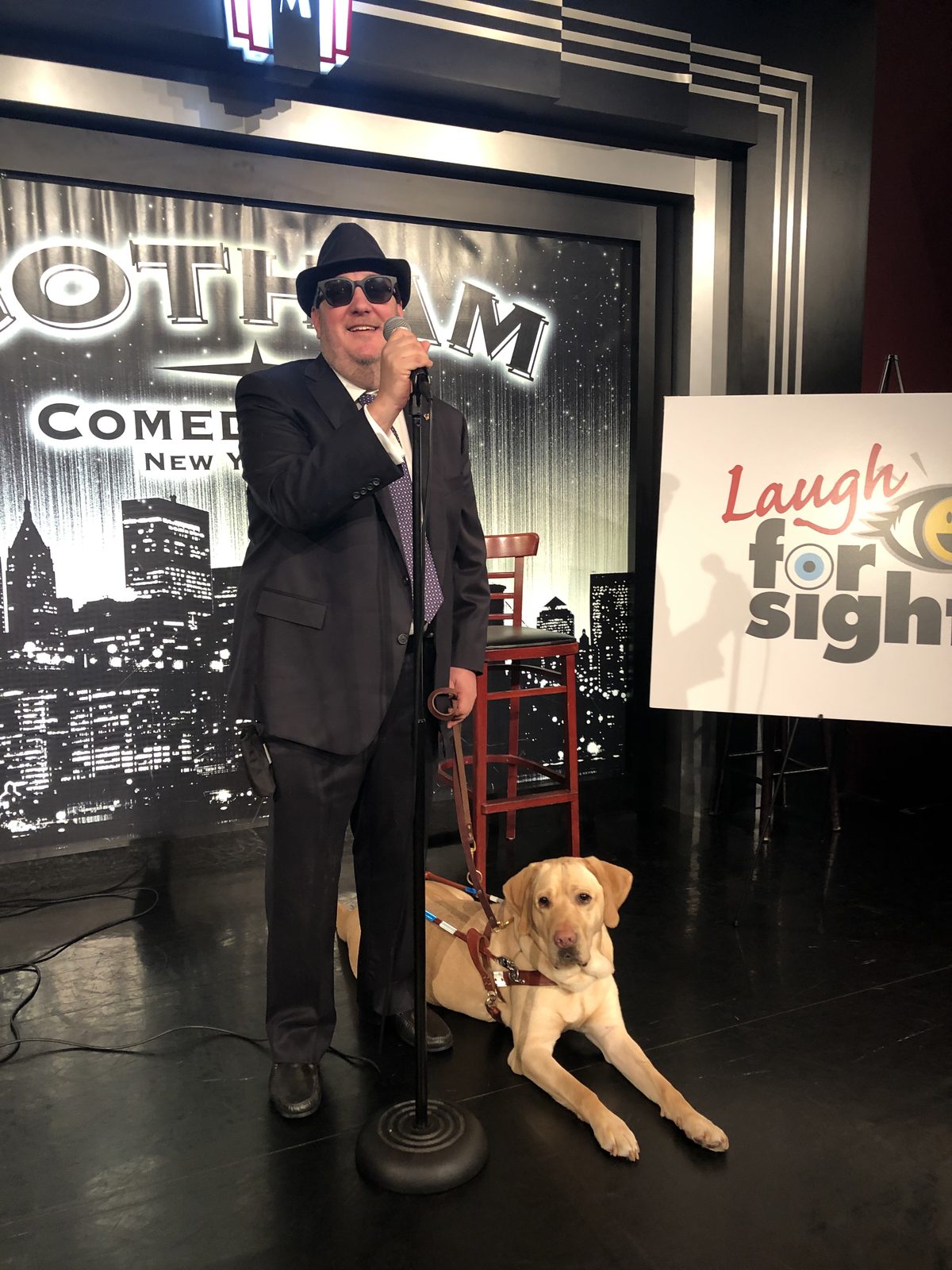 Brian Fischler on stage at Gotham Comedy Club in a dark suit and fedora, holding a microphone and smiling, with his yellow Labrador guide dog Wesley lying in harness at his feet beside a Laugh For Sight sign.