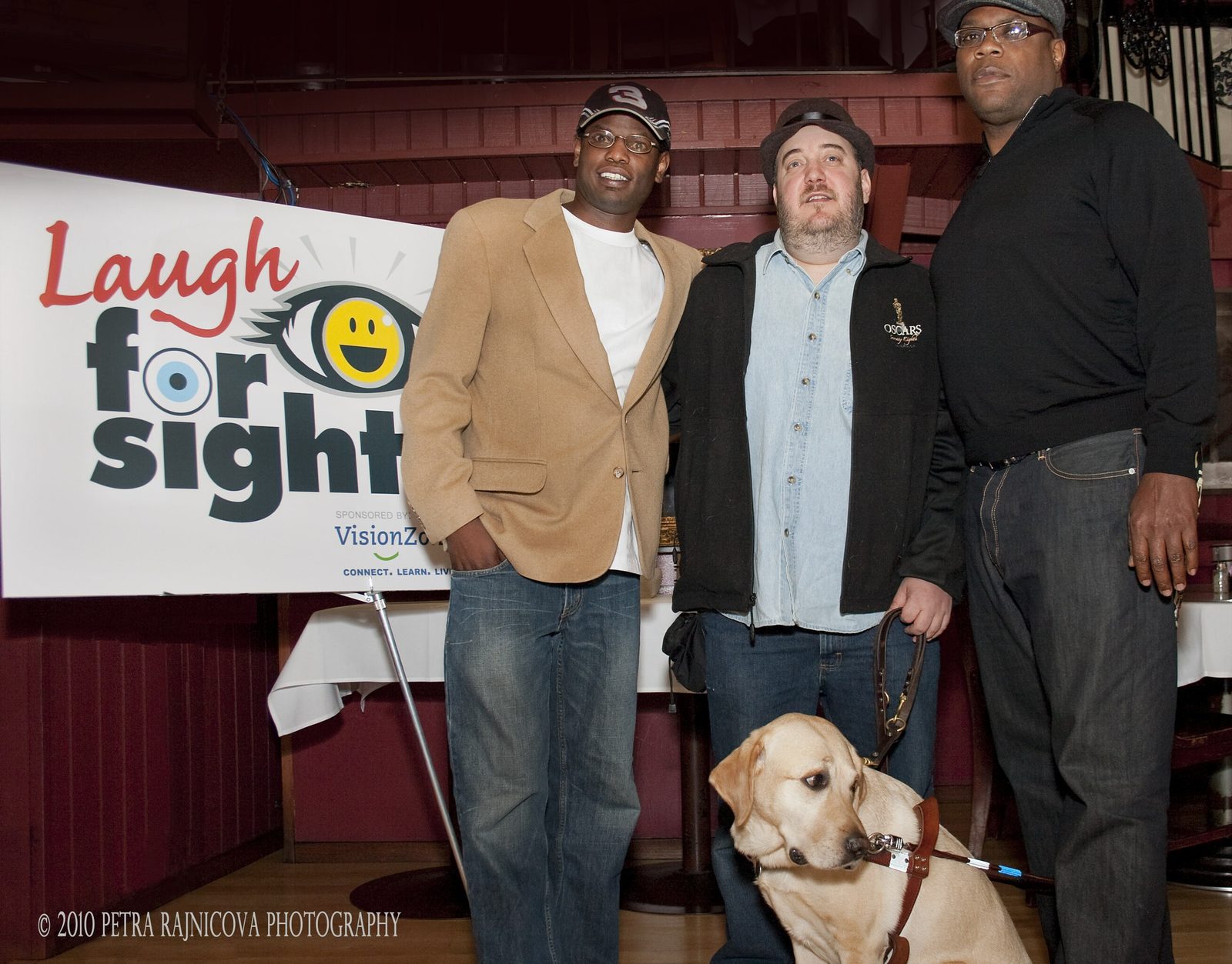 Three men standing together beside a large Laugh For Sight banner, its logo showing a smiling eye and a line reading sponsored by VisionZone. On the left, Daryl Wright, in a tan blazer over a white T-shirt, a dark baseball cap and wire-framed glasses. In the center, Brian Fischler in a dark flat cap and a black jacket embroidered with an Oscars statuette over a denim shirt, holding a guide dog harness. On the right, Alonzo Bodden, in a black long-sleeved top, a gray flat cap and glasses. Brian's yellow Labrador guide dog Nash sits in harness at their feet, looking off to the side.