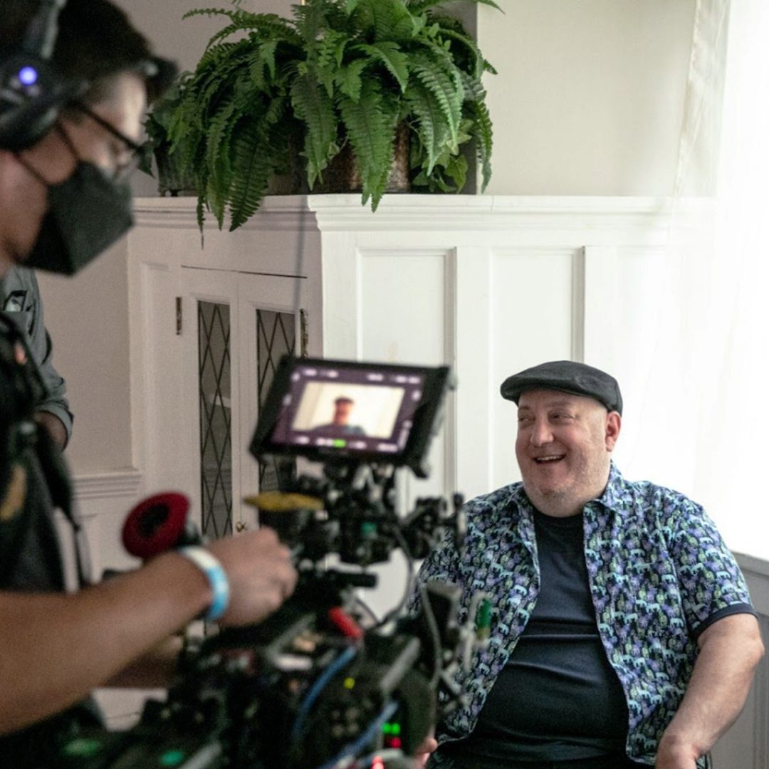 Brian Fischler seated in a bright room with white paneled walls and a hanging fern, laughing mid-conversation. In the foreground a masked camera operator works a cinema camera, Brian's face visible on its monitor. He wears a black flat cap and a zebra-print shirt over a navy tee.
