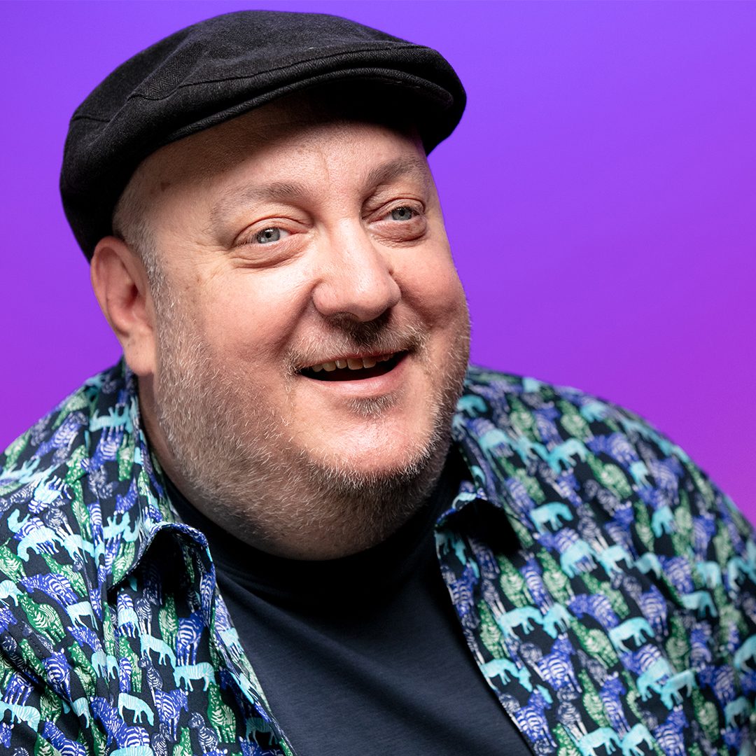 Close portrait of Brian Fischler against a bright purple backdrop, laughing with his head turned slightly to one side. He wears a black flat cap and an open short-sleeved shirt printed with blue and green zebras over a navy tee.