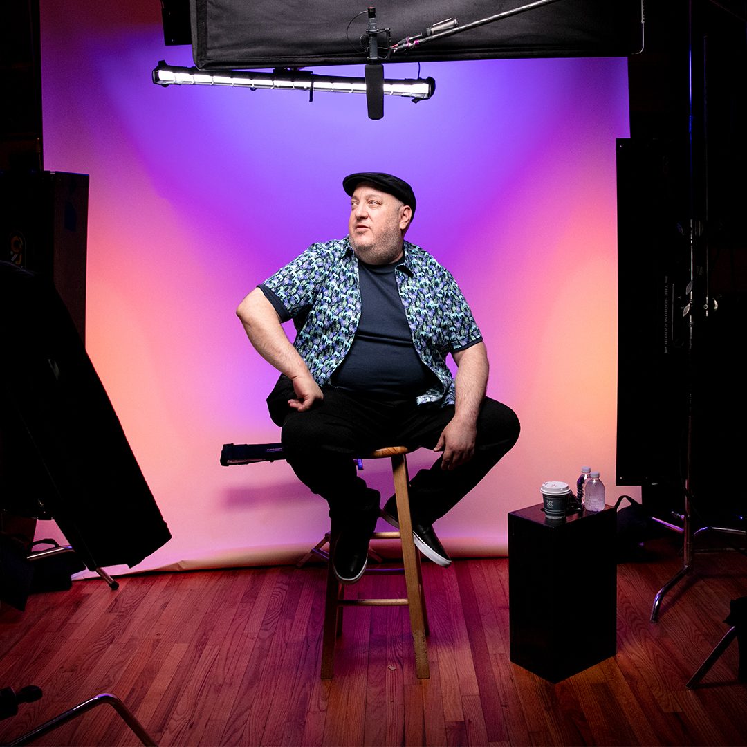 Brian Fischler seated on a wooden stool in the middle of a lit studio set, against a backdrop washed in purple and orange. A softbox and a boom microphone hang overhead, flags and light stands crowd both edges of the frame, and a coffee cup and water bottles sit on a black box beside him. He wears a black flat cap, a zebra-print shirt over a navy tee, and black trousers.