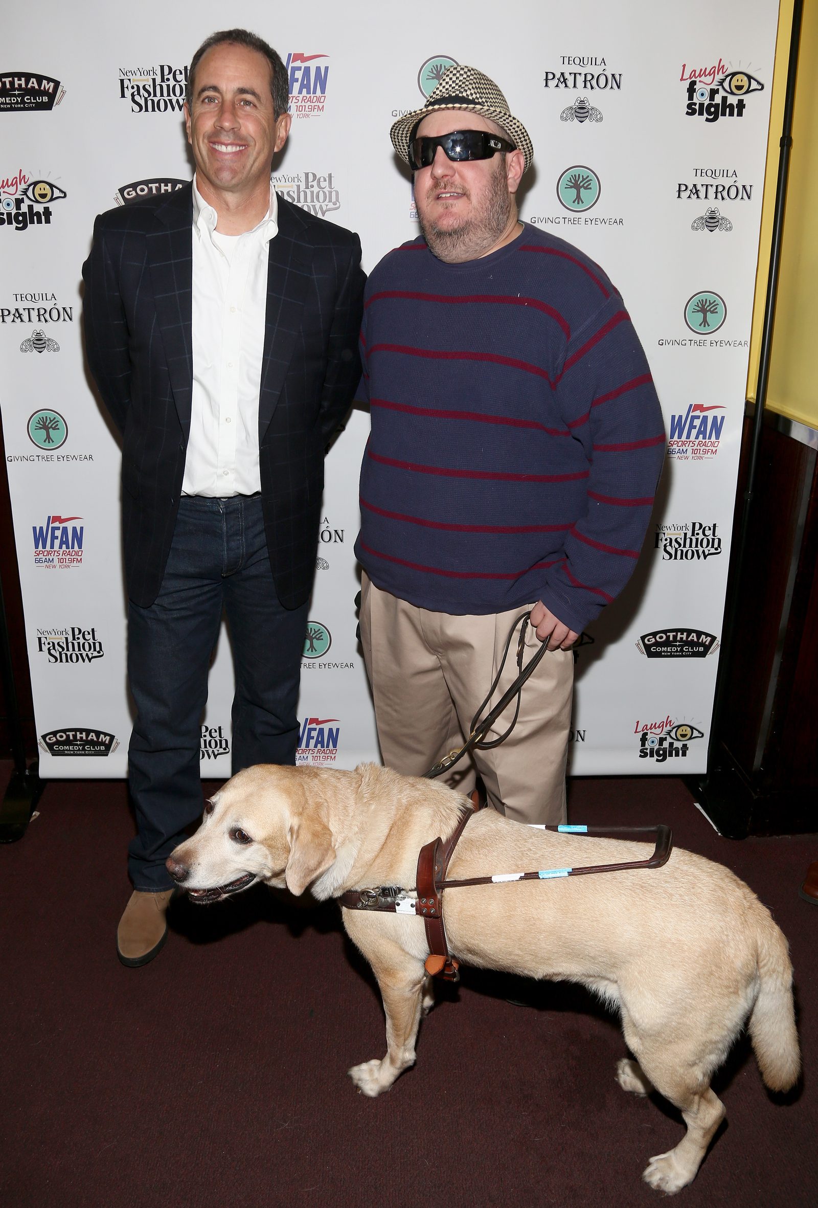 Jerry Seinfeld and Brian Fischler stand together in front of a Laugh For Sight step-and-repeat backdrop. Seinfeld wears a navy blazer over a white shirt and jeans. Brian wears a straw fedora, dark sunglasses, and a navy sweater with red stripes, holding the leash of his guide dog Nash, a yellow Labrador in a leather harness standing across the front of the frame.