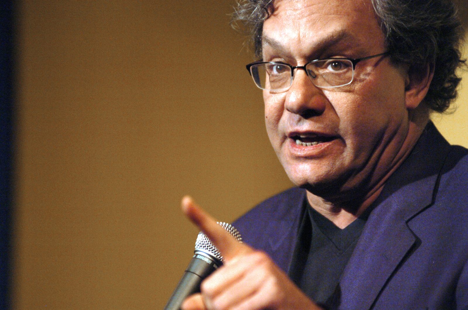 Close-up of Lewis Black on stage at Gotham Comedy Club during the inaugural Laugh For Sight. He holds a microphone in one hand and points out toward the audience with the other, mid-sentence, wearing a blue blazer over a dark shirt.