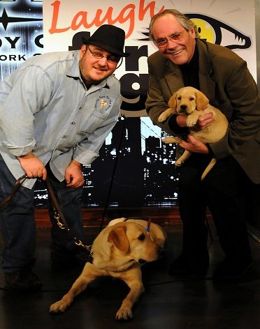 Brian Fischler with his guide dog Nash, a yellow Labrador lying at his feet, standing beside comedian Robert Klein, who cradles a Guiding Eyes puppy in training in his arms. The Laugh For Sight banner hangs behind them at Gotham Comedy Club.